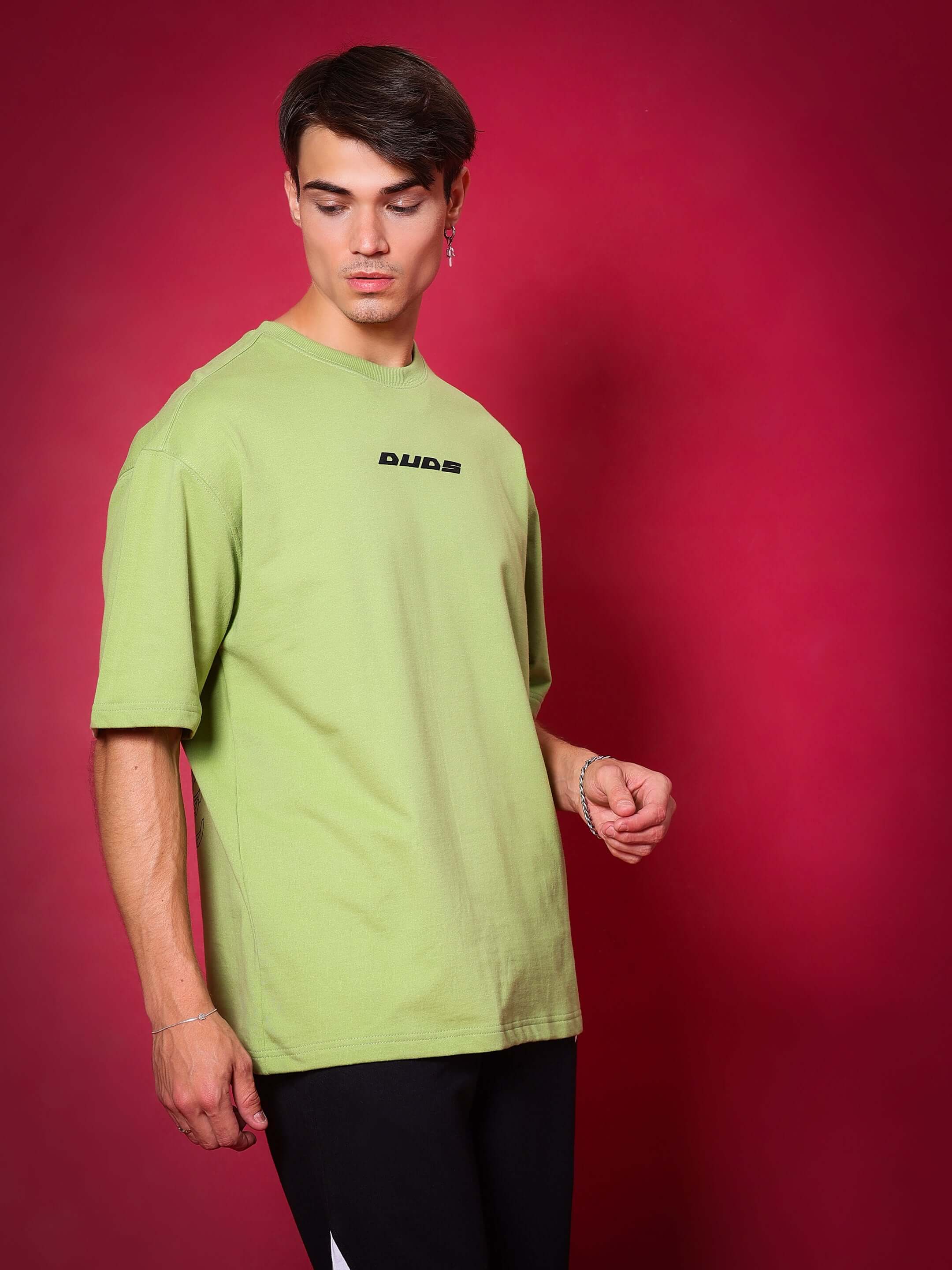 Model wearing SCORPIO over-sized t-shirt in pista green, made from premium cotton fabric, standing against a red backdrop.