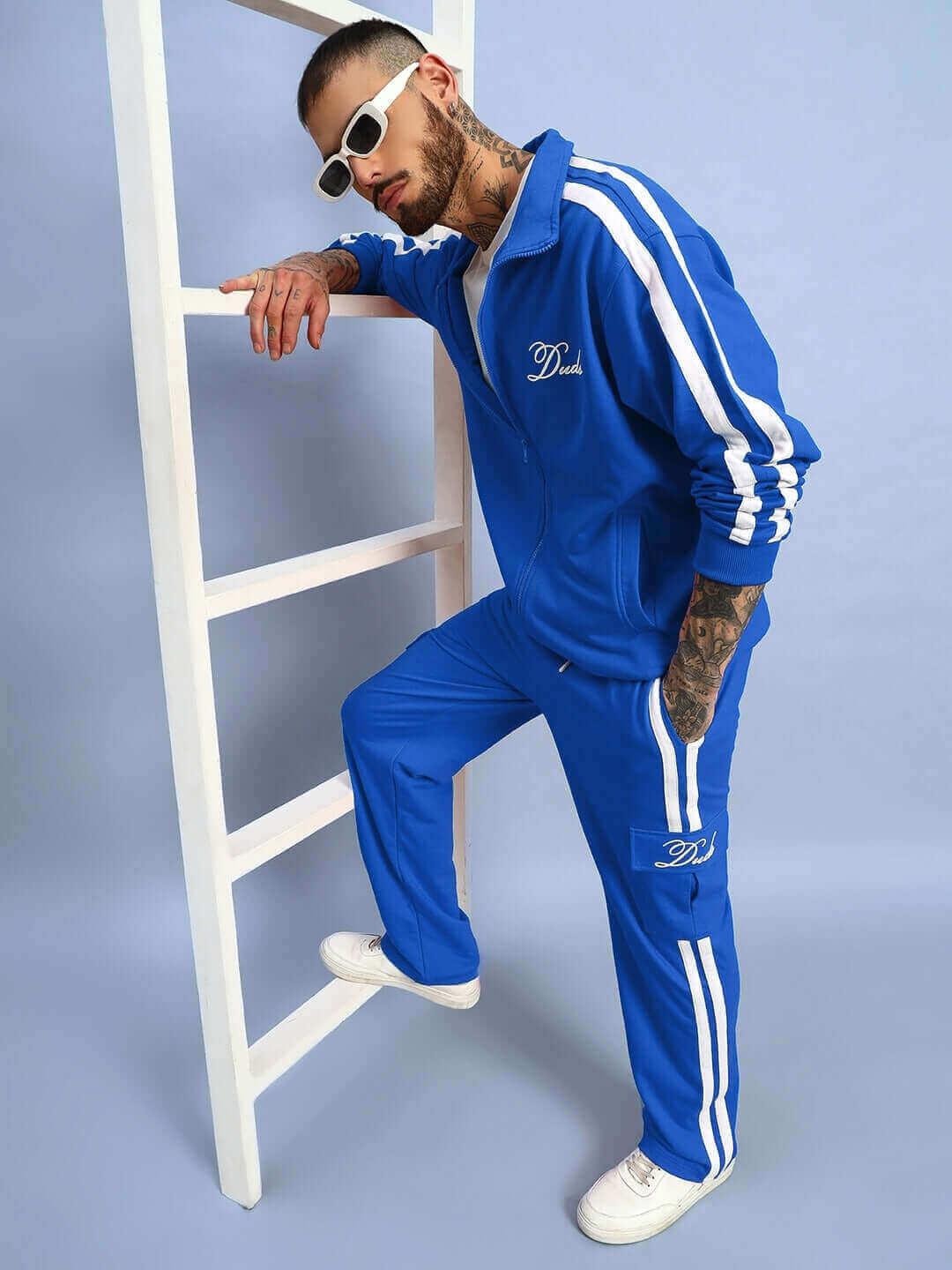 Model wearing Scotia Jacket in royal blue with white stripes, leaning on a ladder, showcasing sporty style for DUDS brand.