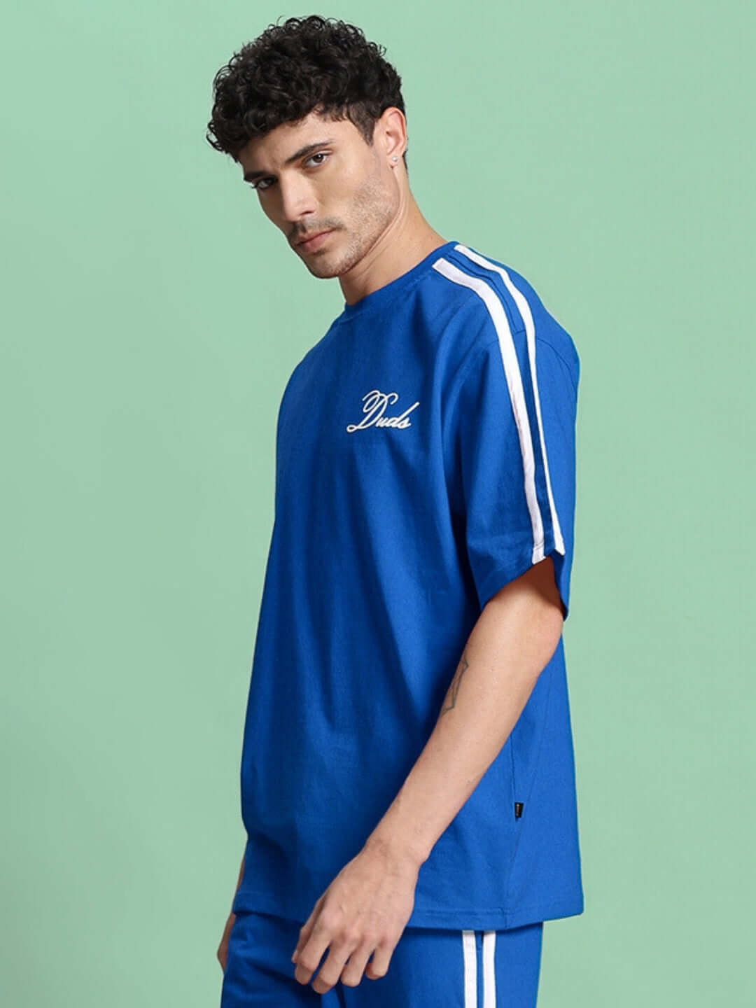 SCOTIA OVER-SIZED T-SHIRT BLUE in premium heavy gauge fabric with white stripes on the shoulder, modeled against a green background.