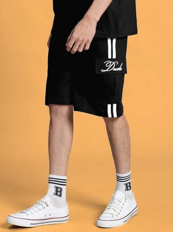 Model wearing SCOTIA REGULAR FIT SHORTS BLACK with white sneakers, showcasing casual summer style on yellow background.