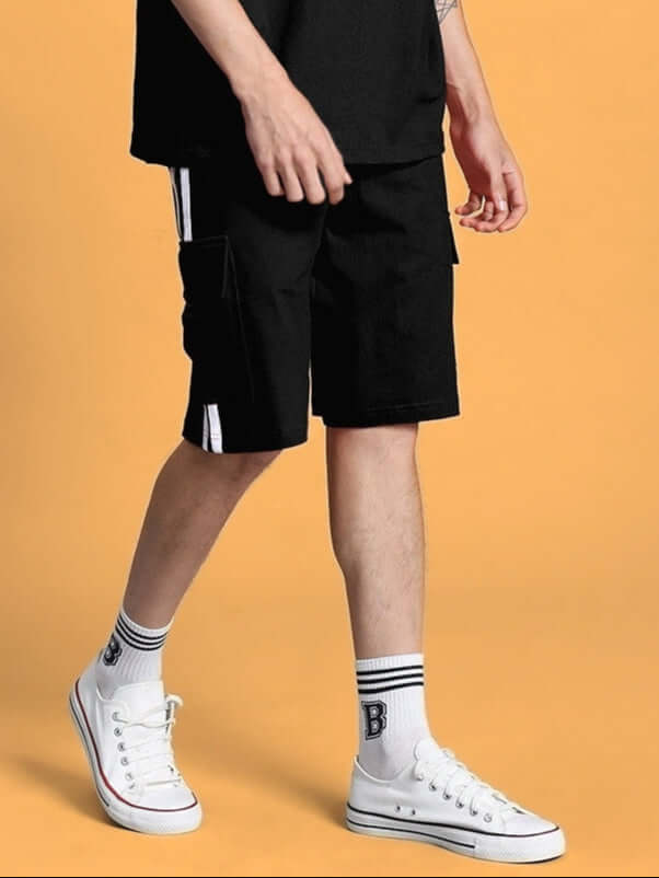 SCOTIA REGULAR FIT SHORTS BLACK worn by a model standing in white sneakers on a yellow background, showcasing casual style.