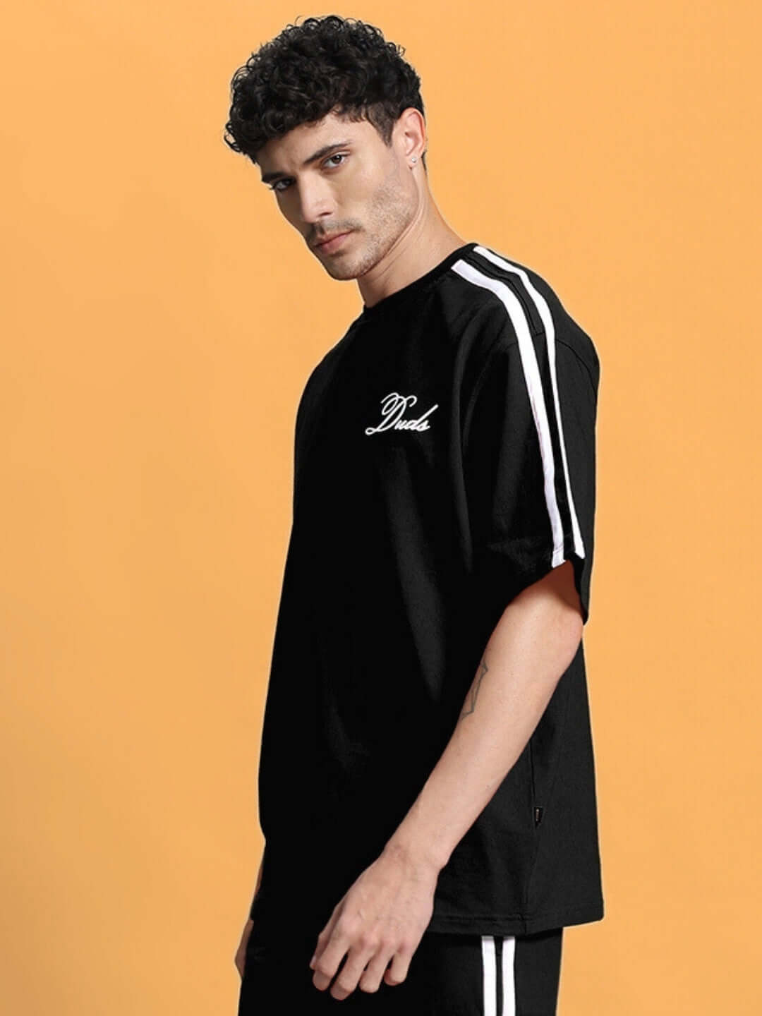Man wearing SCOTIA OVER-SIZED T-SHIRT BLACK with stripe details on the sleeve against orange background.