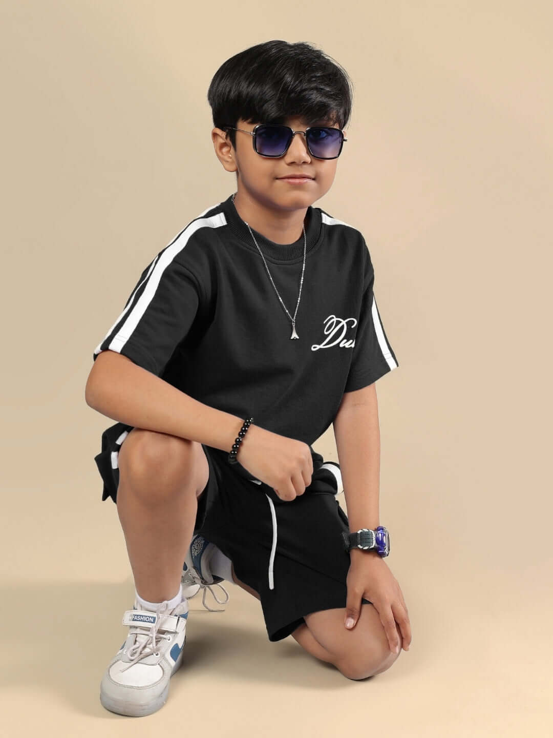 Child wearing SCOTIA SHORTS FOR BOYS & GIRLS BLACK, sitting on the floor in casual pose with sunglasses.