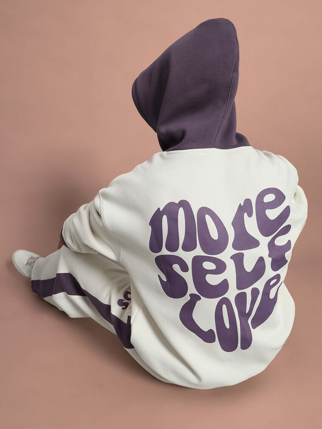 Back view of SELF LOVE FLEECE OVERSIZED HOODIE WHITE VIOLET featuring 'More Self Love' graphic on a seated model with hood up.