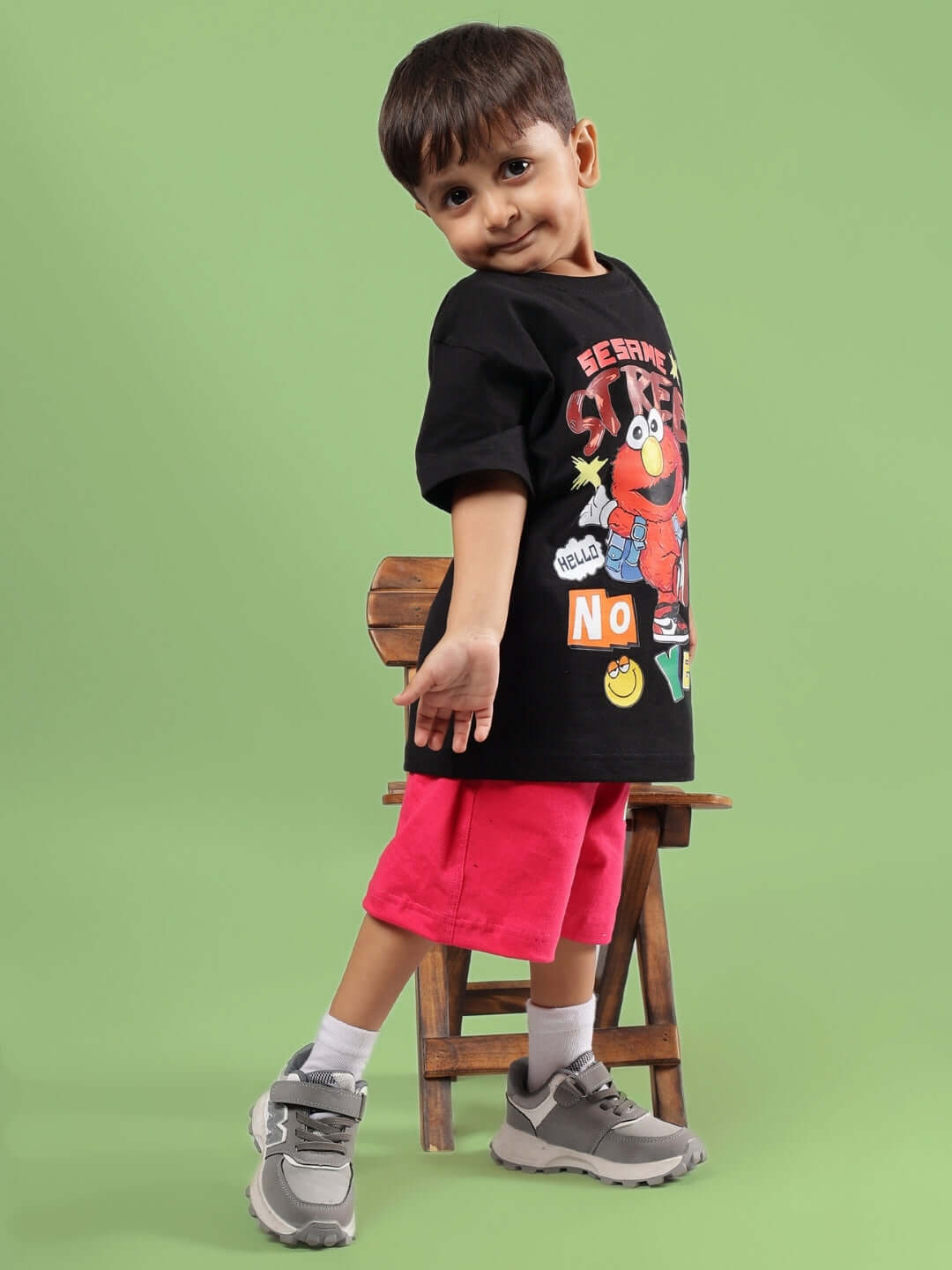 Child wearing Sesame T-Shirt for Boys Girls Black, standing in front of a green background, styled with pink shorts and sneakers.
