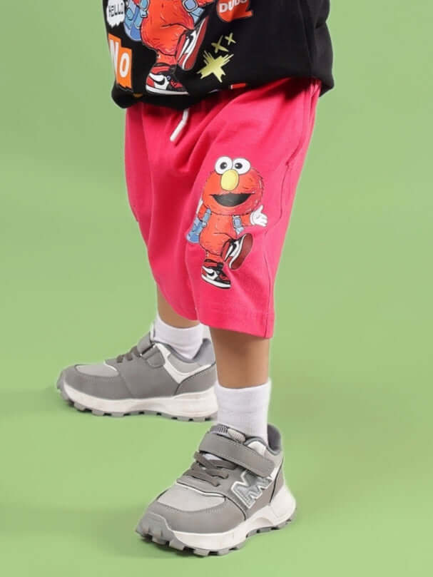 Child wearing SESAME SHORTS FOR BOYS & GIRLS PINK with Elmo print, grey sneakers on a green background.