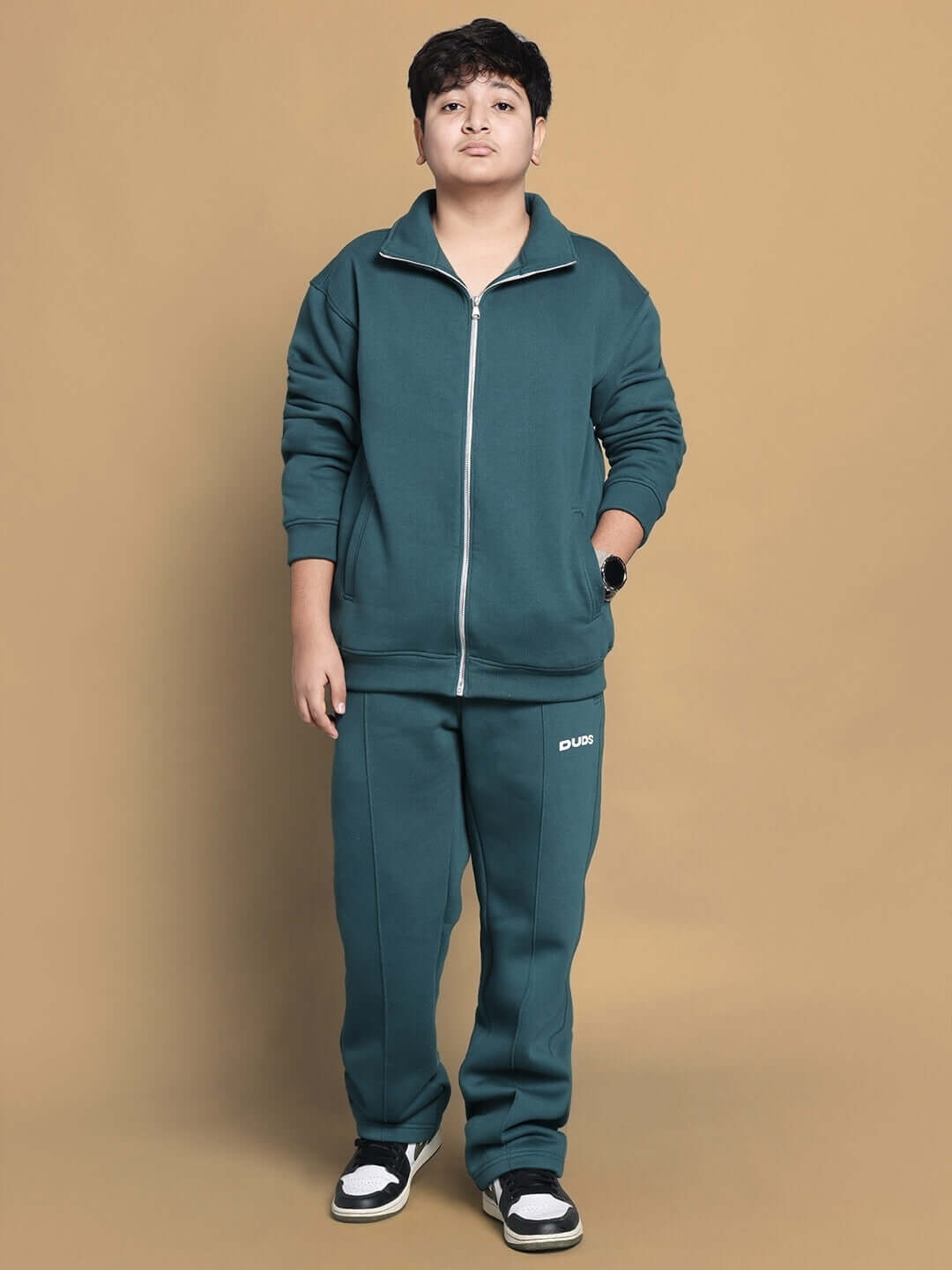 Young person wearing teal green fleece jogger set stands against a tan background.