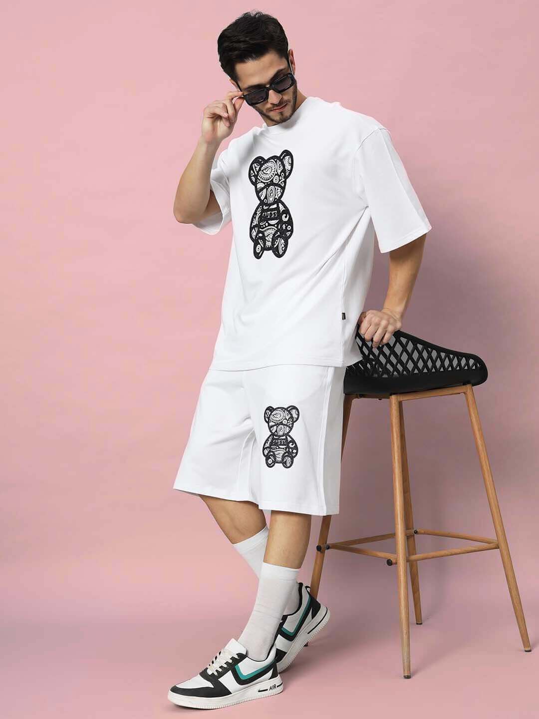 Model wearing SHIMMY over-sized white t-shirt with bear graphic, paired with matching shorts, sunglasses, and sneakers, in pink backdrop.