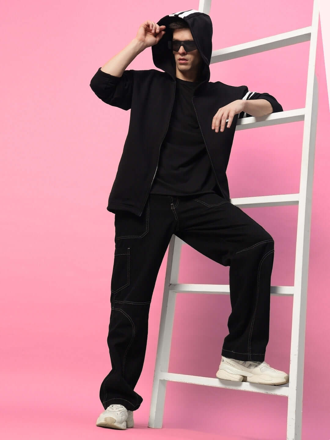 Model in SHINOBI ZIPPER LONG LENGTH FLEECE HOODIE BLACK posing against a pink backdrop, highlighting its sleek, modern streetwear design.