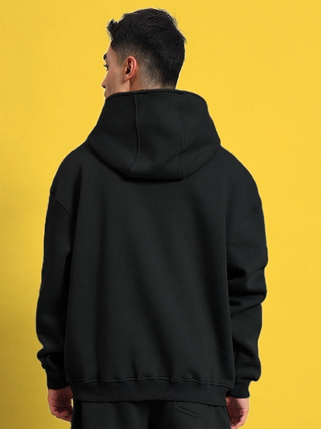 Man wearing black Shooter cotton fleece hoodie, back view against yellow background. Made-to-order by DUDS tailors with custom fit option.