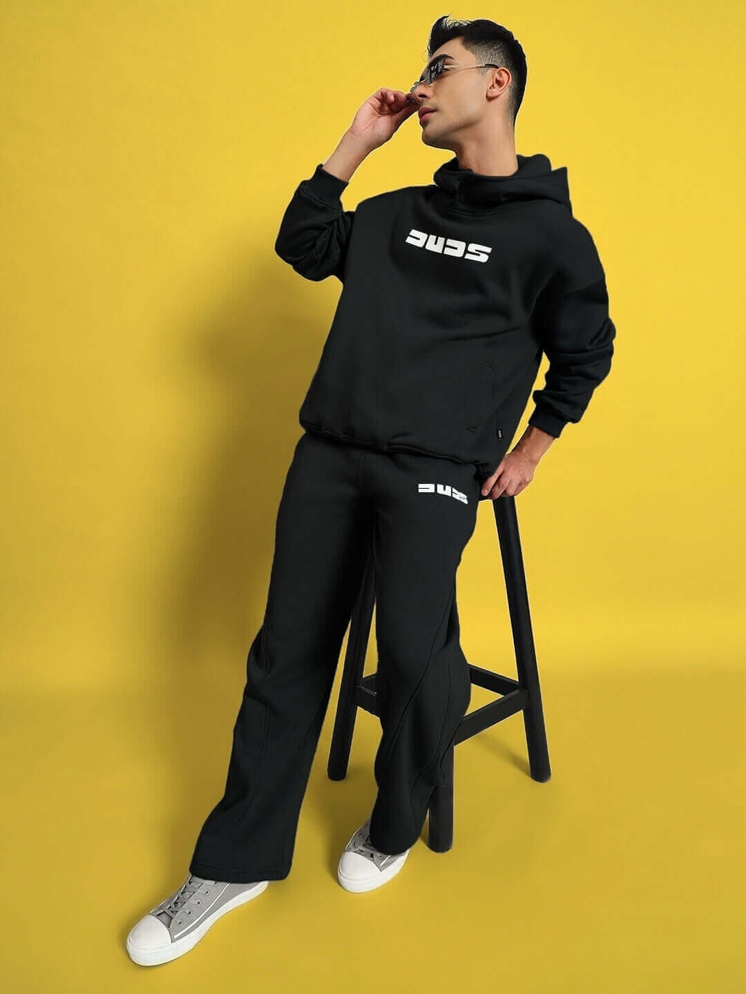 Model wearing WEARDUDS SHOOTER FLEECE CO-ORD BLACK with sneaker shoes and glasses, standing against a yellow background.