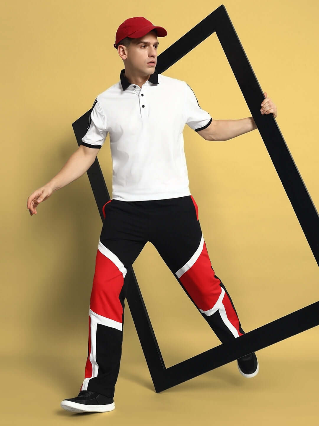 Model wearing SHROUD Relaxed Fit Joggers in black and red, paired with a white polo and red cap, holding a geometric frame.