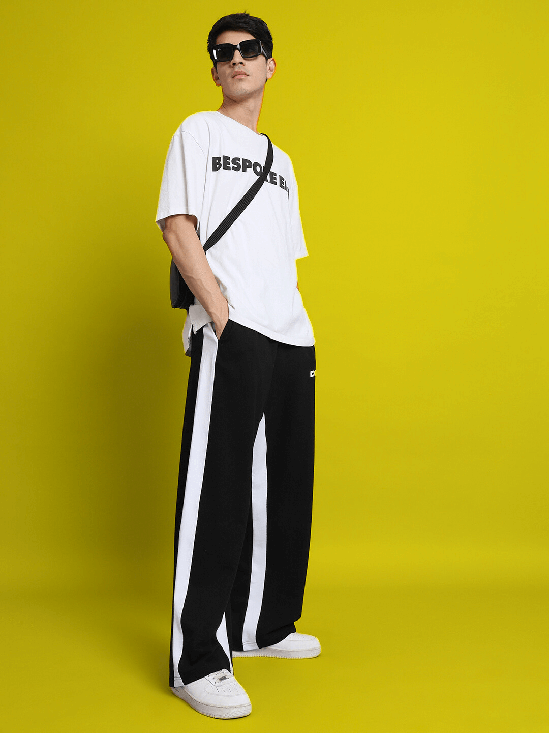 Model wearing black side seam baggy joggers by DUDS, styled with a white t-shirt and sunglasses against a yellow background.