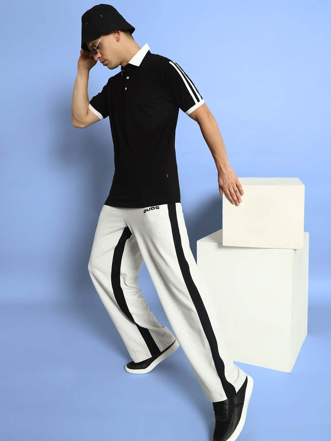 Model wearing SIDE SEAM BAGGY JOGGERS in melange grey-black from DUDS, styled with a black polo shirt and cap, posing on blue background.