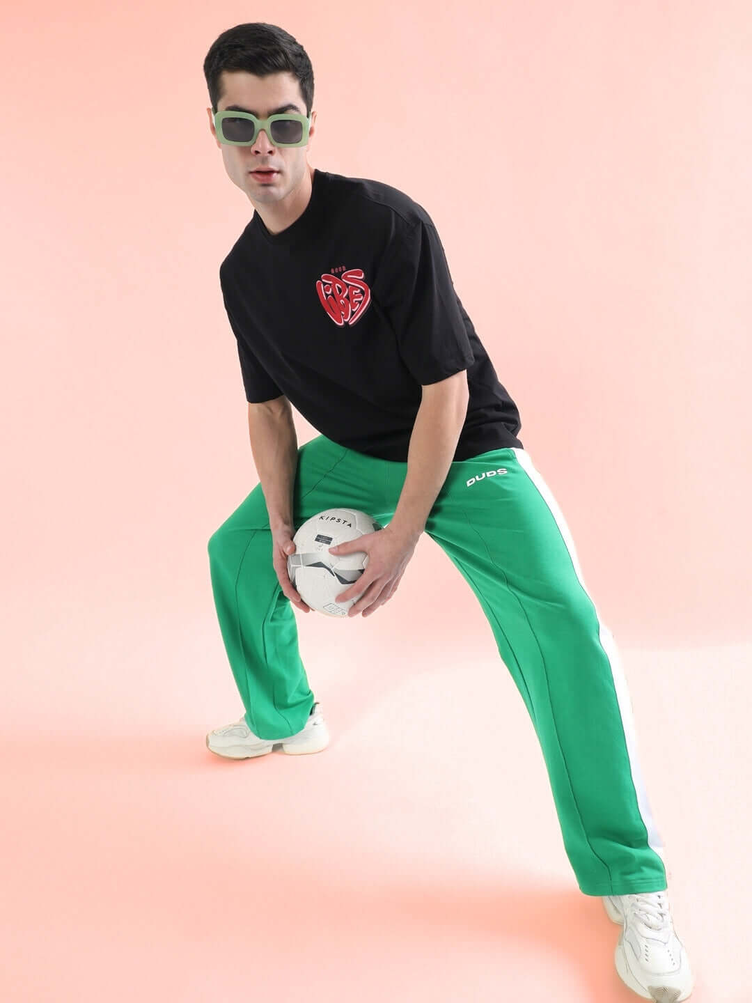 Model wearing SIDE SEAM PLATED JOGGERS in green, posing with a soccer ball against a pink background, highlighting sporty and stylish appeal.