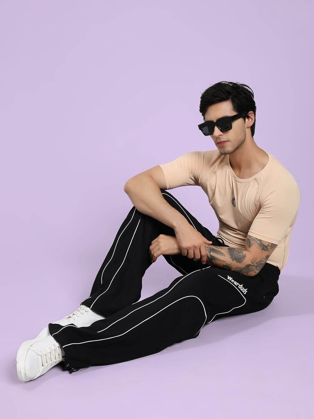 Model wearing SIGNATURE PIPING JOGGERS BLACK with contrast piping, sitting stylishly against a purple backdrop, showcasing casual fashion.
