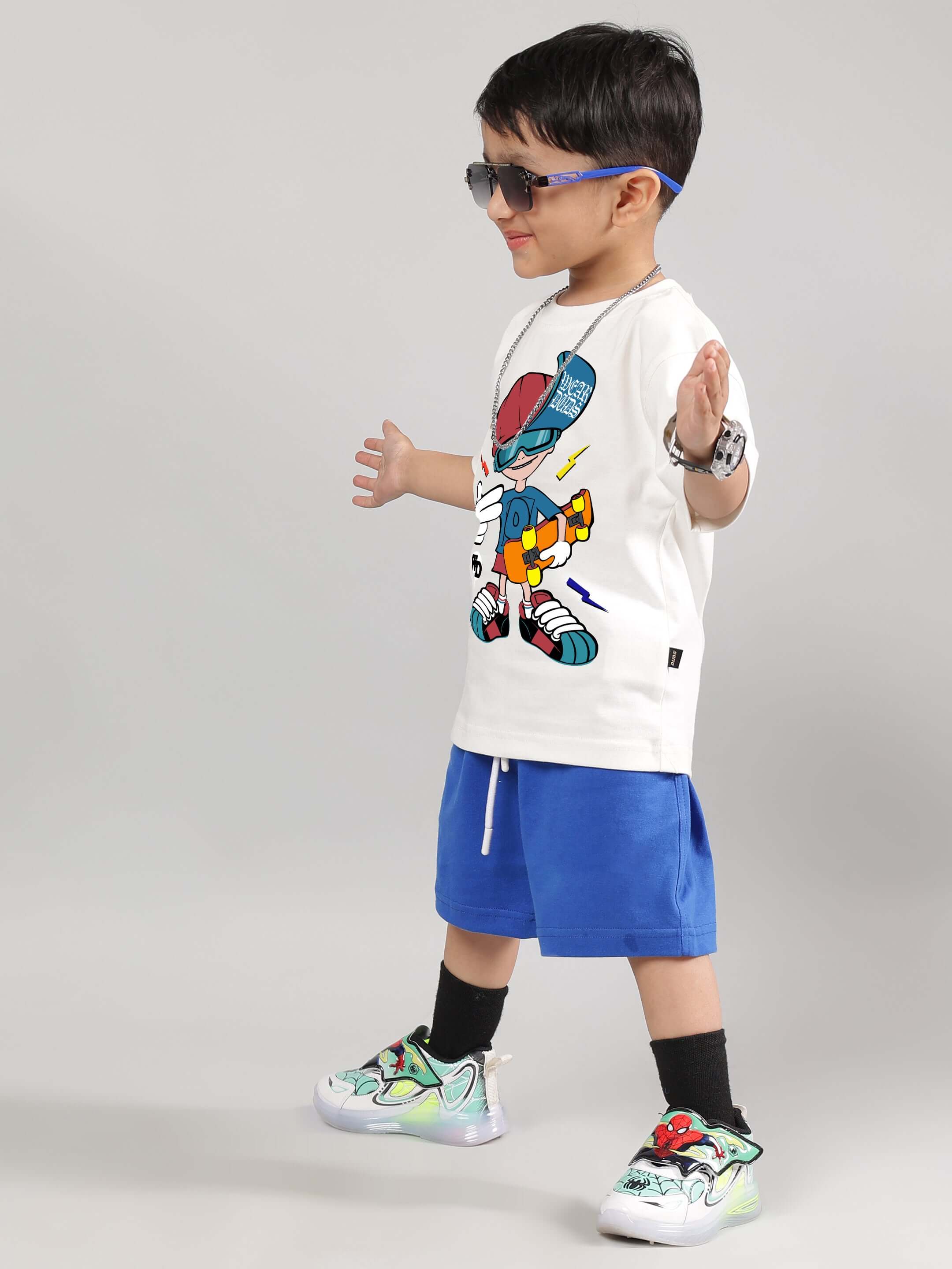 Child wearing off-white SKATE RIDER T-Shirt featured for boys and girls, styled with blue shorts and colorful sneakers.