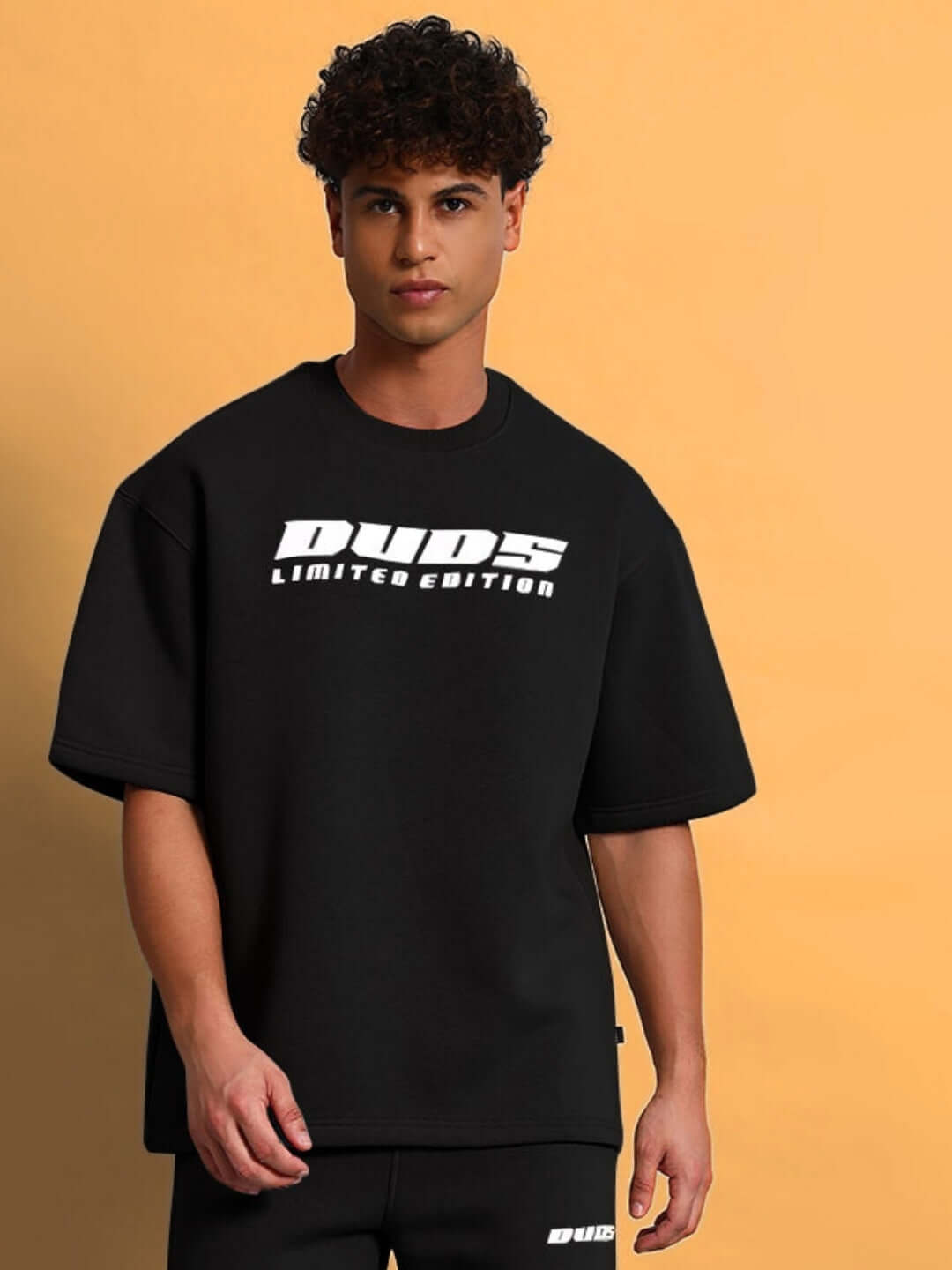 Model wearing SKODA FLEECE OVERSIZED T-SHIRT BLACK with bold logo, showcasing casual streetwear style against a warm background.