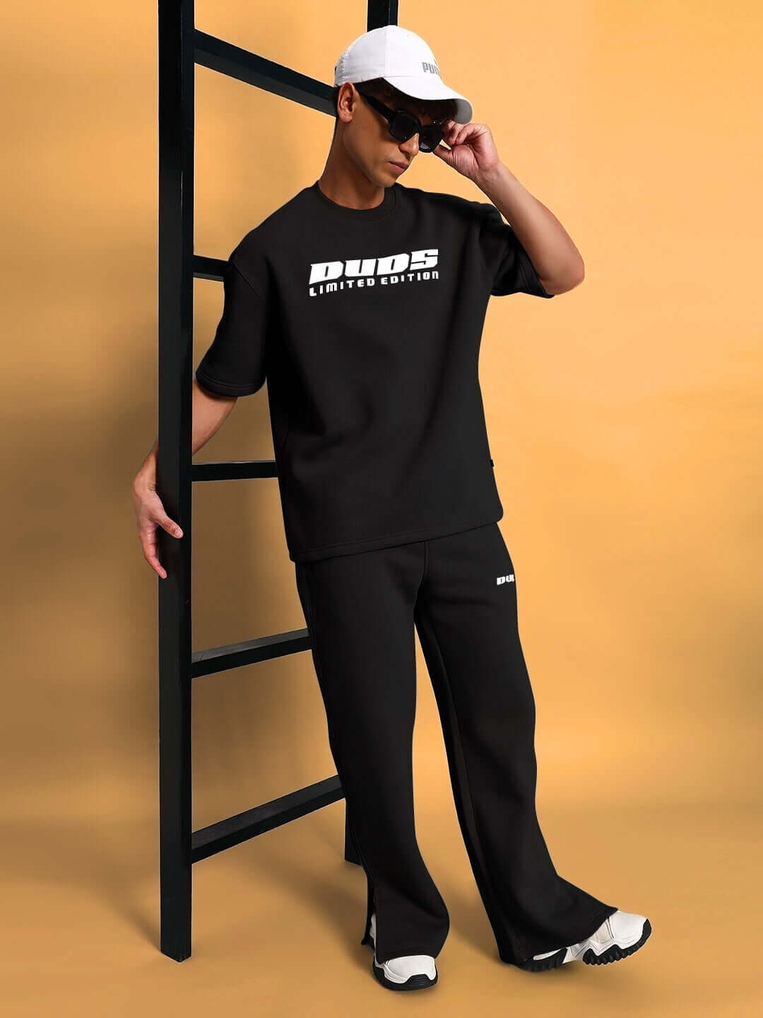 Model wearing WEARDUDS SKODA FLEECE CO-ORD BLACK with cap and sunglasses, standing against ladder.