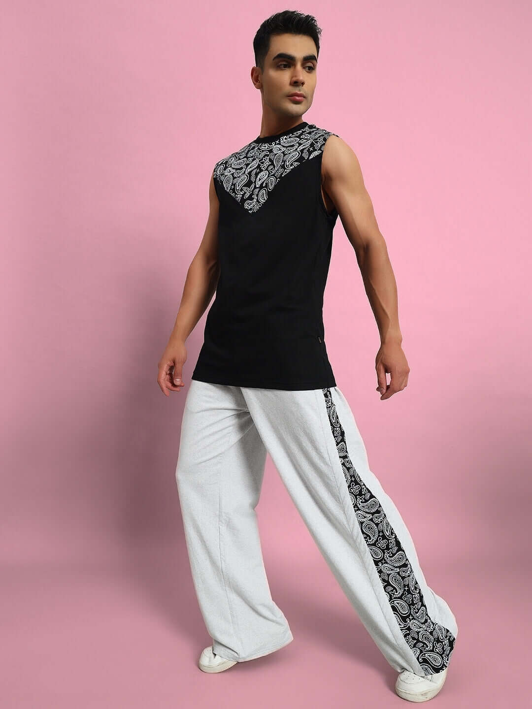 Fashion model wearing black sleeveless top with printed details and white wide-legged pants, standing against a pink background.