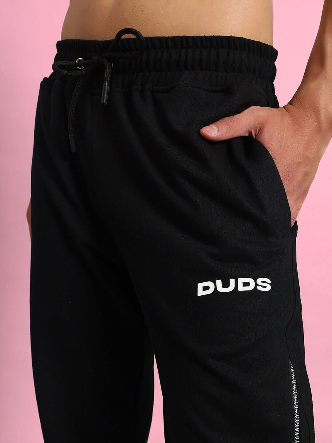 Close-up of model wearing black DUDS Slider Joggers with adjustable drawstring and side pockets against pink background.