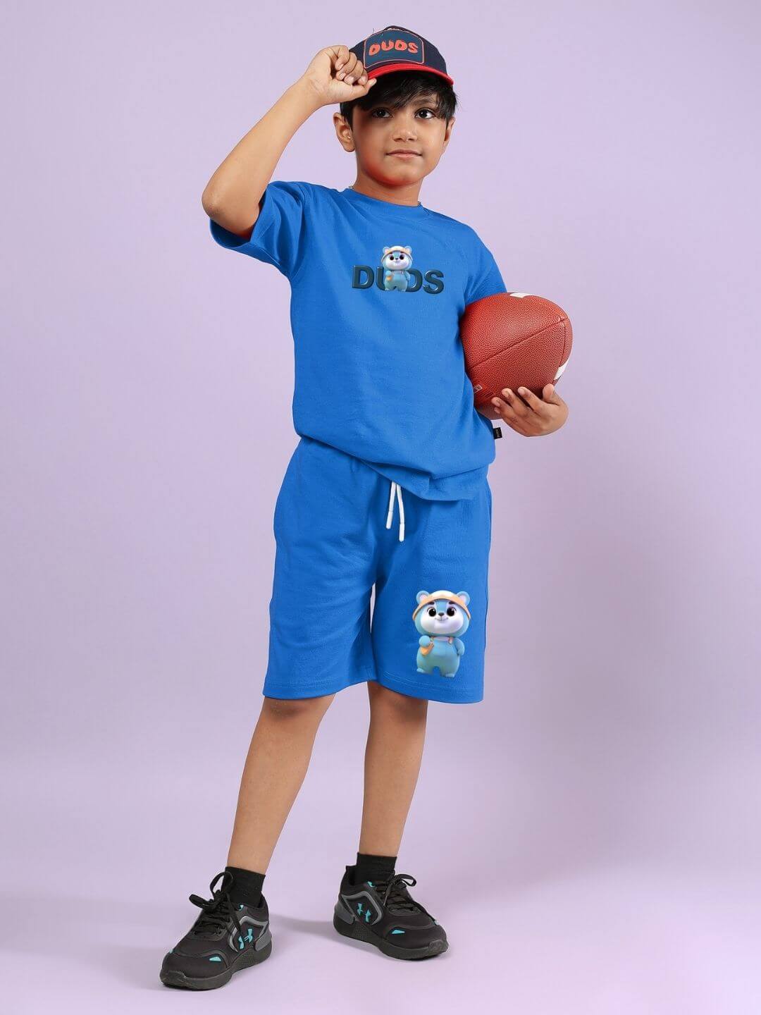 Boy wearing blue Smiley Bear shorts set with cap, holding a football. Comfy unisex summer outfit for active kids.