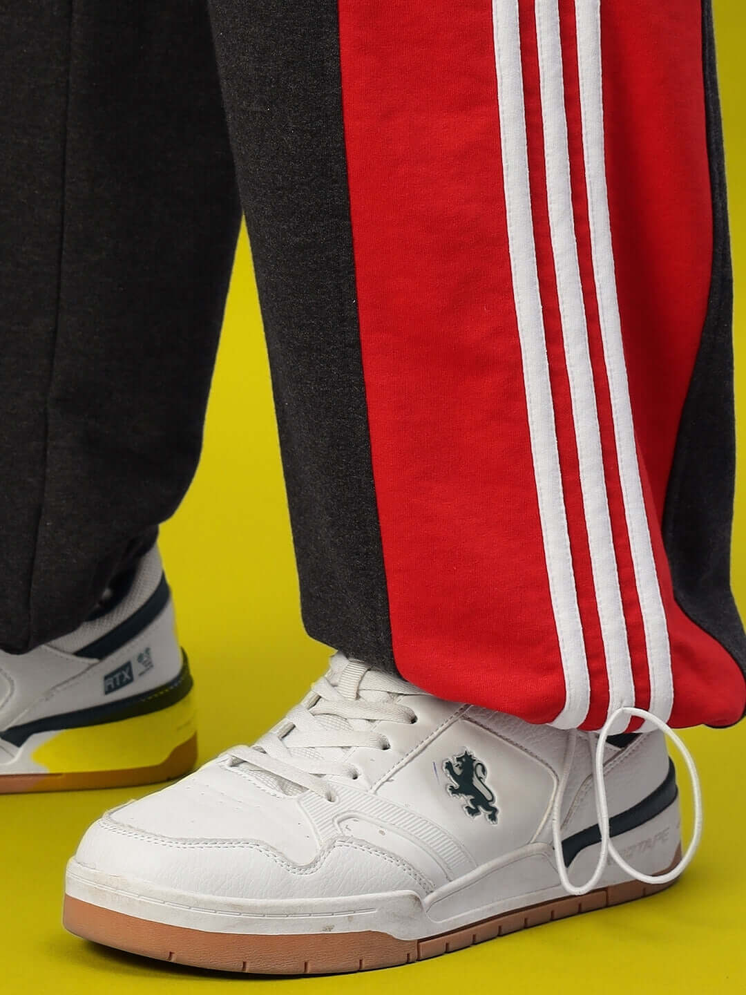 Close-up of SNICKER relaxed-fit joggers in dark grey and red with white stripes paired with white sneakers on a yellow background.
