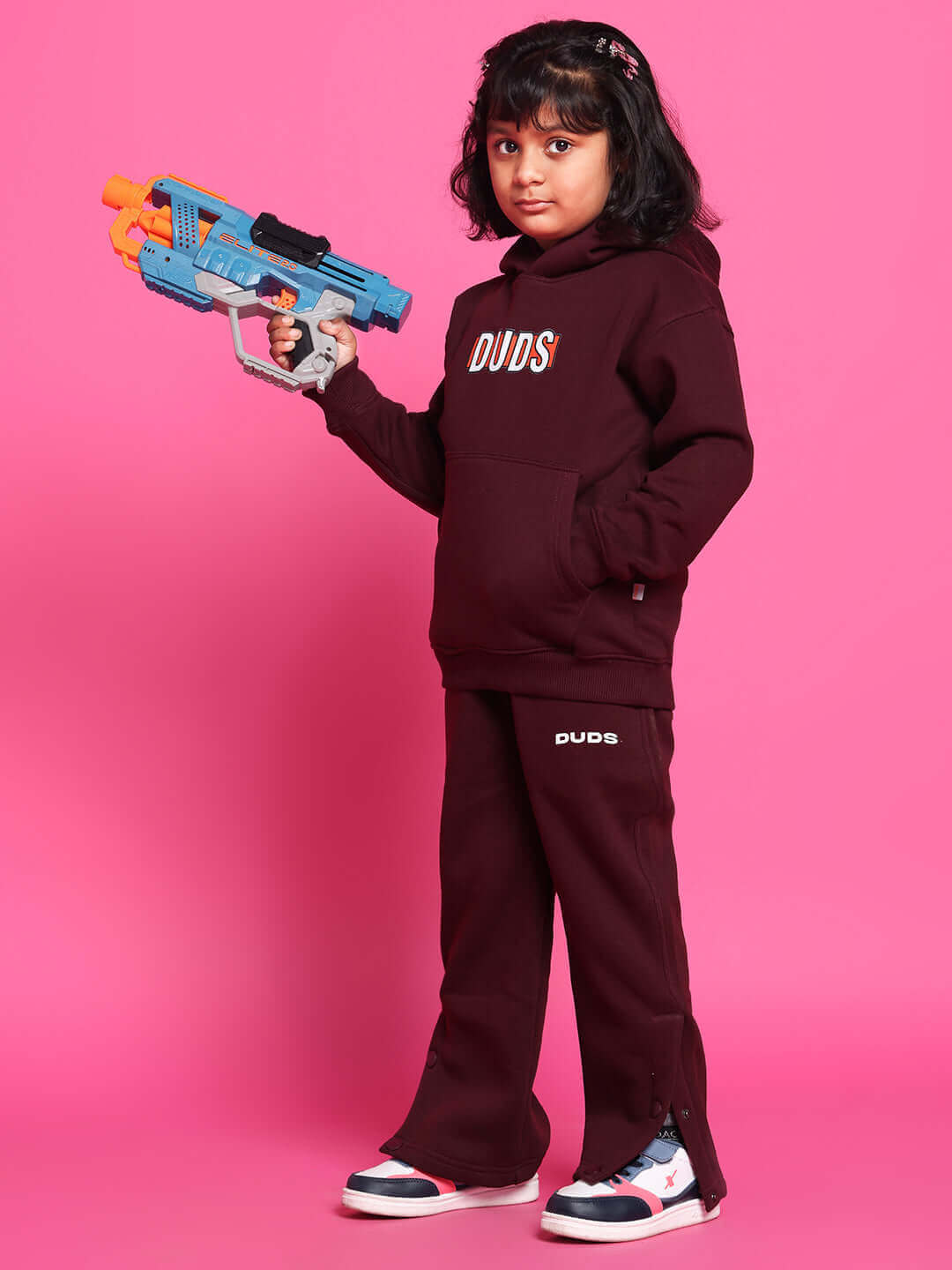Child modeling Snoopy fleece pant and hoodie in wine color, holding a toy gun, against a pink background.