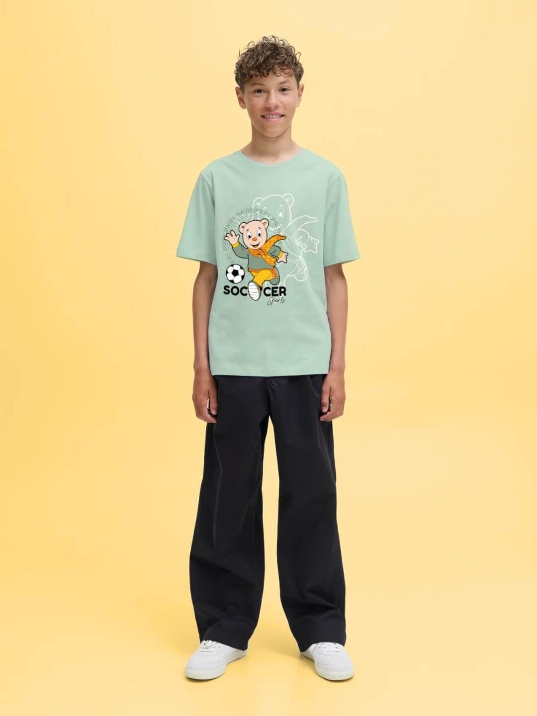 Boy wearing SOCCER PRINTED T-SHIRT FOR BOYS AND GIRLS LIGHT GREEN with graphic design, standing against yellow background