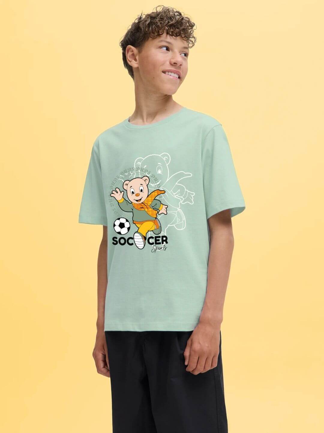 Boy wearing SOCCER PRINTED T-SHIRT FOR BOYS AND GIRLS LIGHT GREEN with playful soccer graphic design against a yellow background.