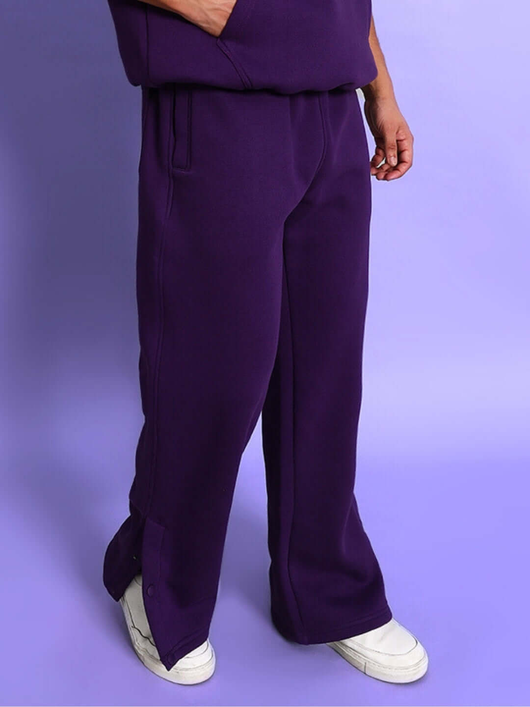 Person wearing Sooty Fleece Pant in purple, showing relaxed fit and side buttons, styled with white sneakers on a purple background.