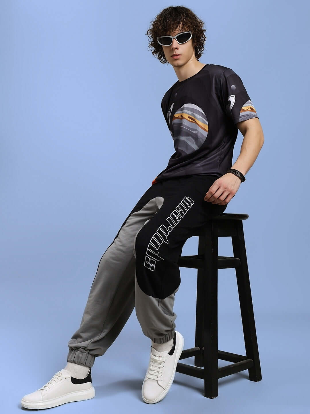 Model wearing SPACE FANTASY black t-shirt, sports polyester, seated on stool, styled with graphic joggers and white sneakers.