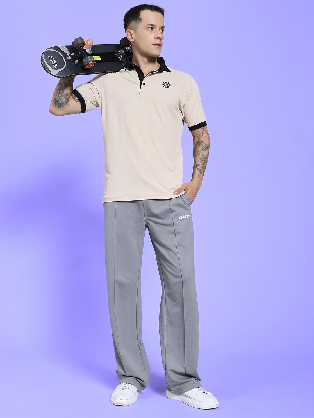 Man in SPIRAL RELAXED-FIT JOGGERS GREY holding a skateboard against a purple background, wearing a light beige polo shirt.