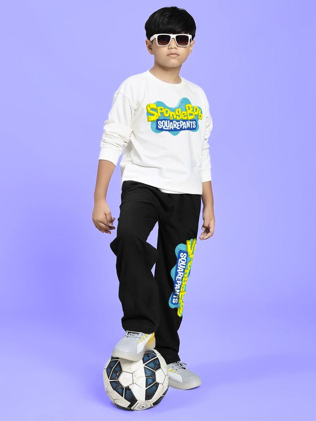Child poses in SPONGEBOB SQUAREPANTS CO-ORD FOR BOYS AND GIRLS WHITE BLACK with soccer ball, wearing sunglasses, on a purple background.