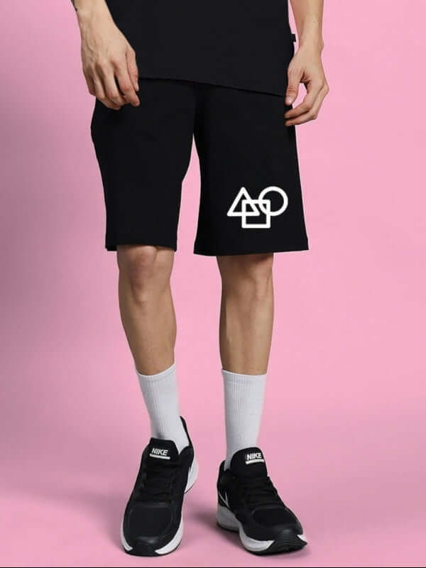 Man wearing SQUID GAME SUMMER SHORTS BLACK with geometric design, standing against pink background