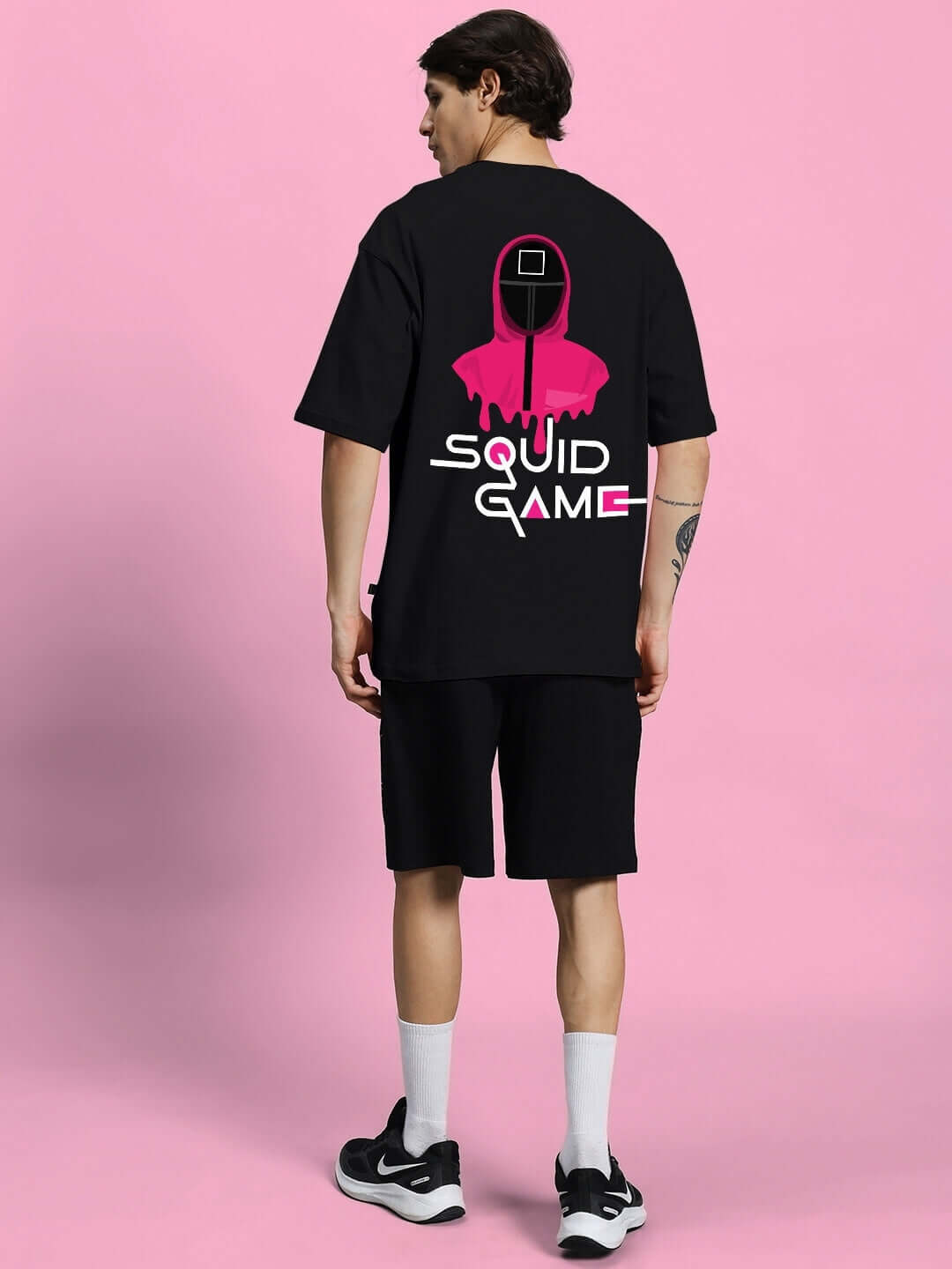 Man wearing SQUID GAME SUMMER CO-ORD SET BLACK, featuring T-shirt and shorts with iconic design on pink background.