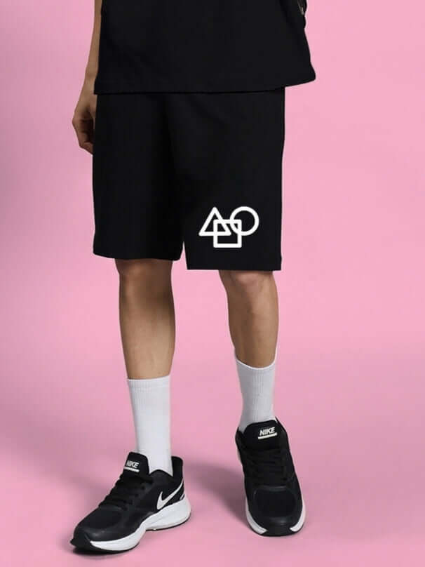 Man wearing SQUID GAME SUMMER SHORTS BLACK with geometric design, displayed against a pink background.