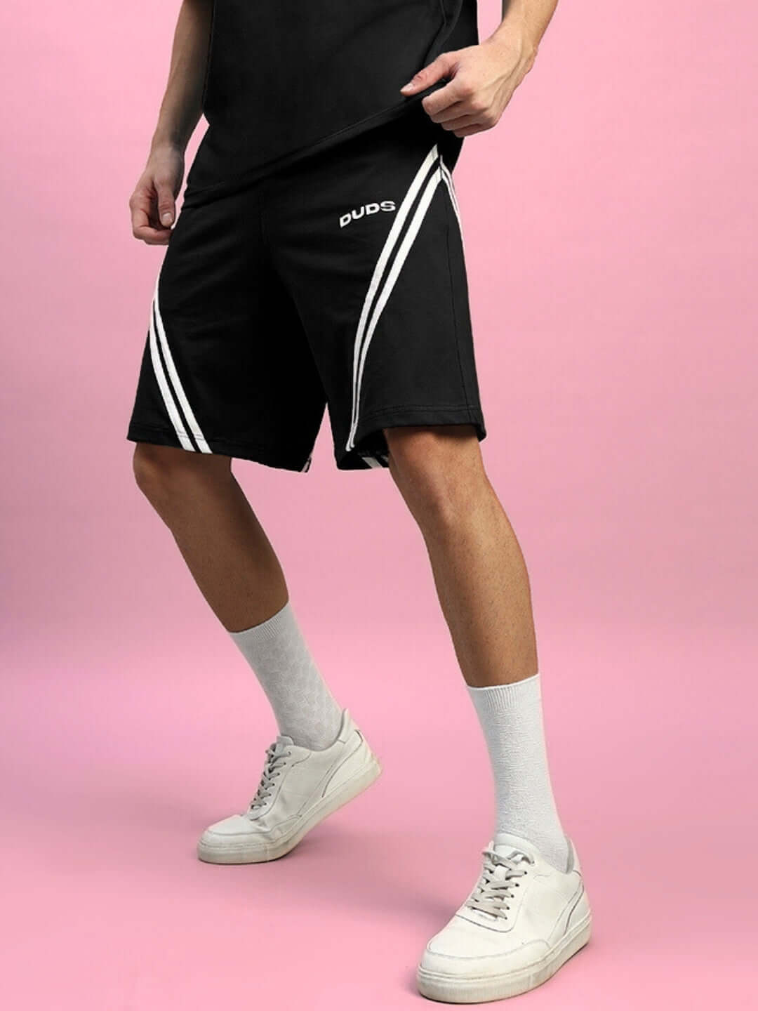 Man wearing DUDS STAR JUMPER regular fit black shorts with white stripes, on a pink background, showcasing casual sporty style.