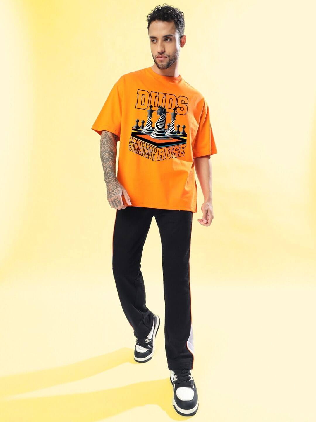 Man wearing STRATEGY RUSE OVER-SIZED T-SHIRT ORANGE with bold graphic, styled in streetwear fashion, posed confidently on yellow background.