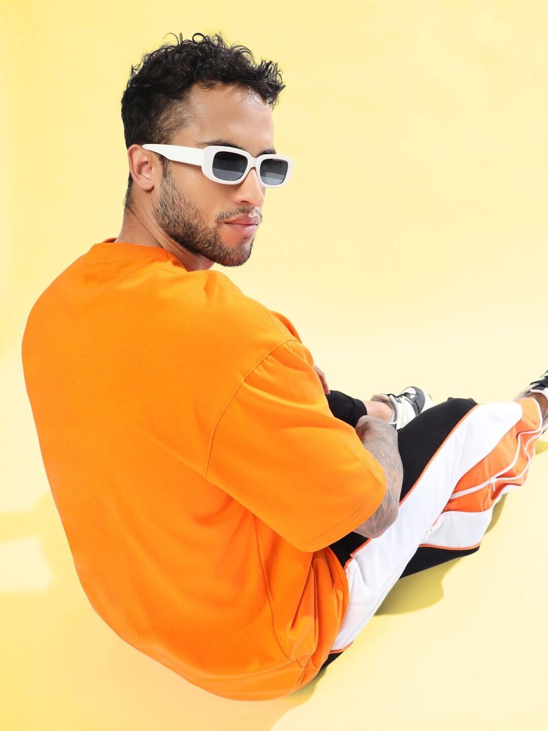 Man wearing STRATEGY RUSE OVER-SIZED T-SHIRT ORANGE with sunglasses seated against a yellow background.
