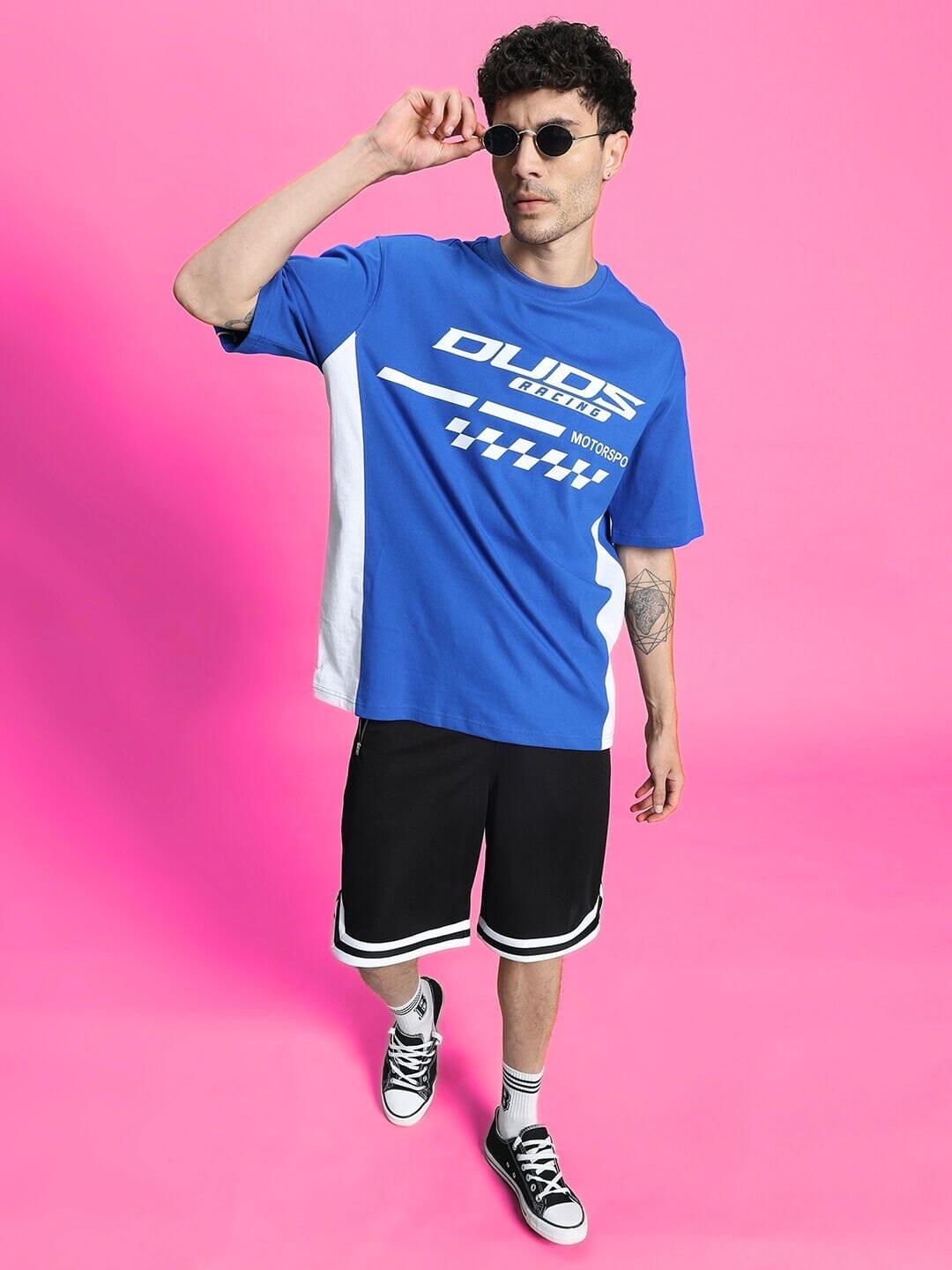 Man wearing Streak Regular Fit Black Shorts with blue graphic tee against pink background, showcasing casual style.