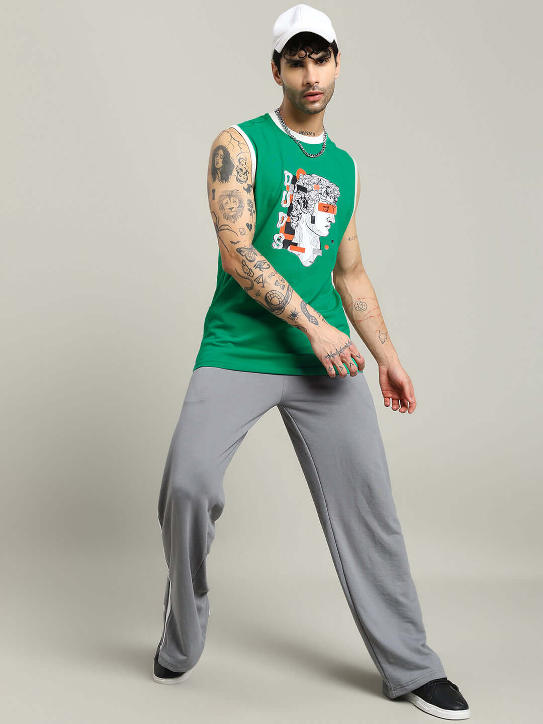 Person wearing STREAM REGULAR FIT T-SHIRT GREEN with tattoos, baseball cap, gray pants, and black shoes, showcasing urban style.