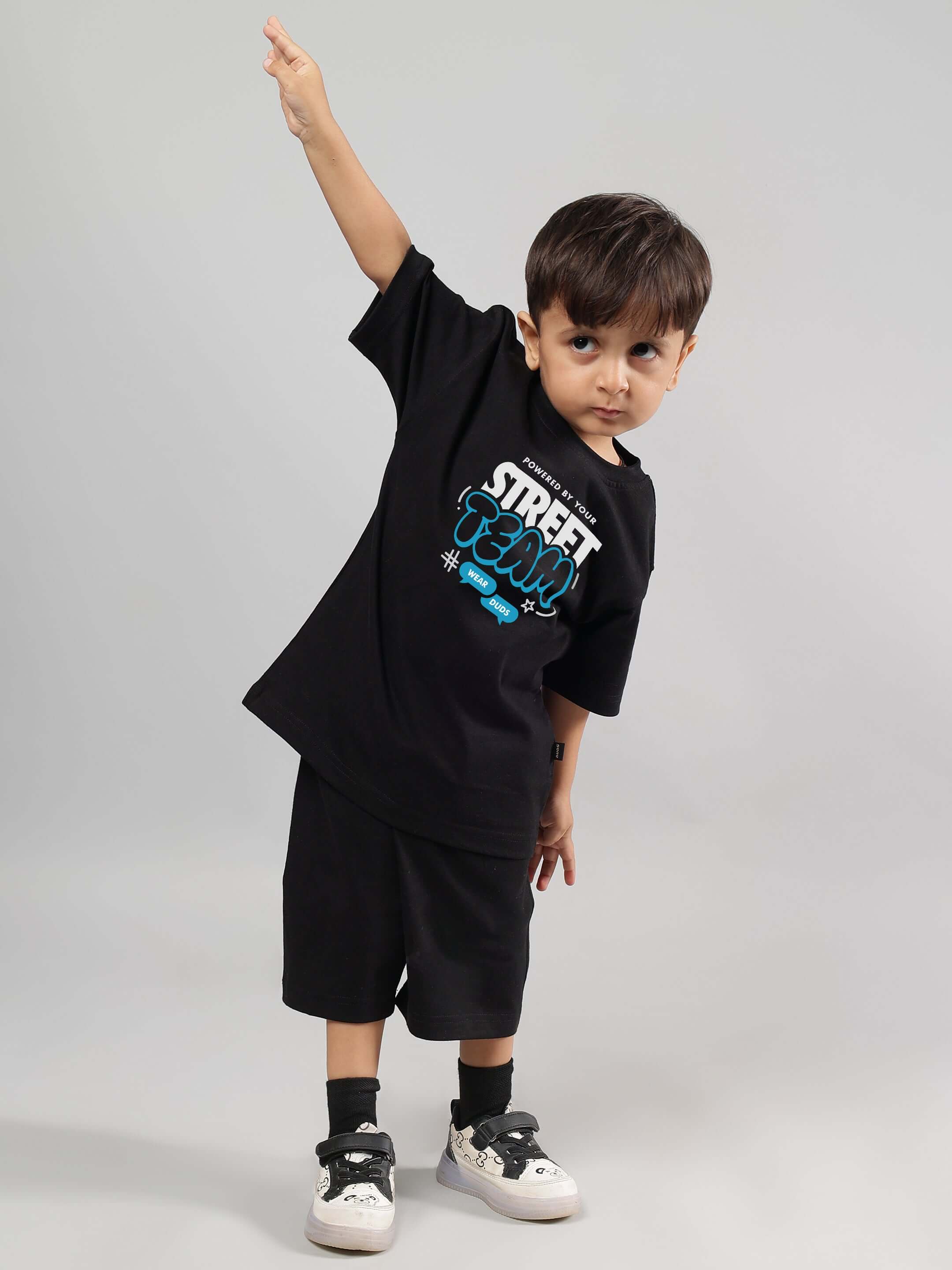 Young boy wearing black "Street Team" coordinated outfit, standing with arm raised in a playful pose. Fashion by DUDS.