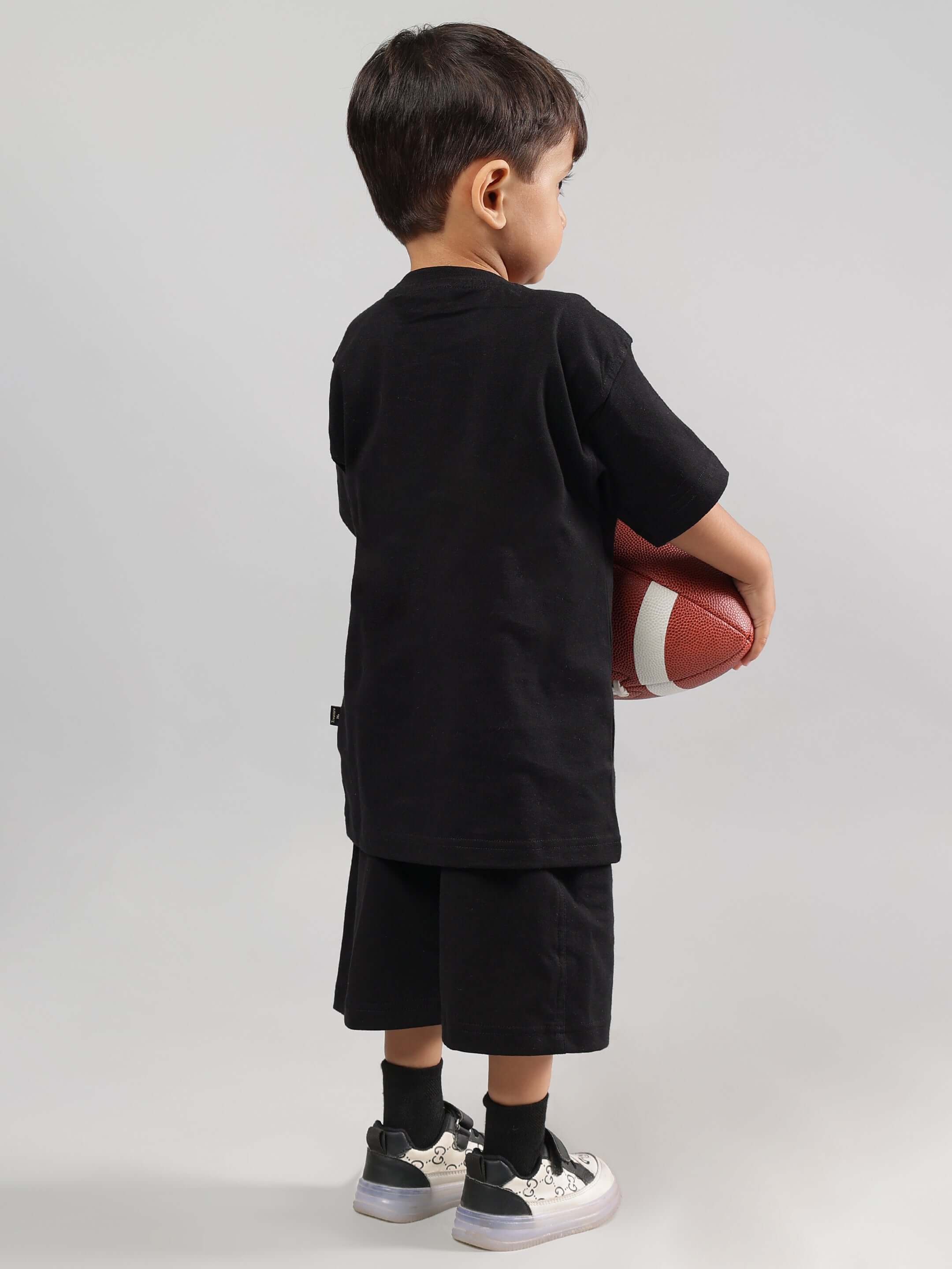 Boy wearing STREET TEAM black t-shirt and shorts, holding a football, showing rear view for boys & girls fashion by DUDS.