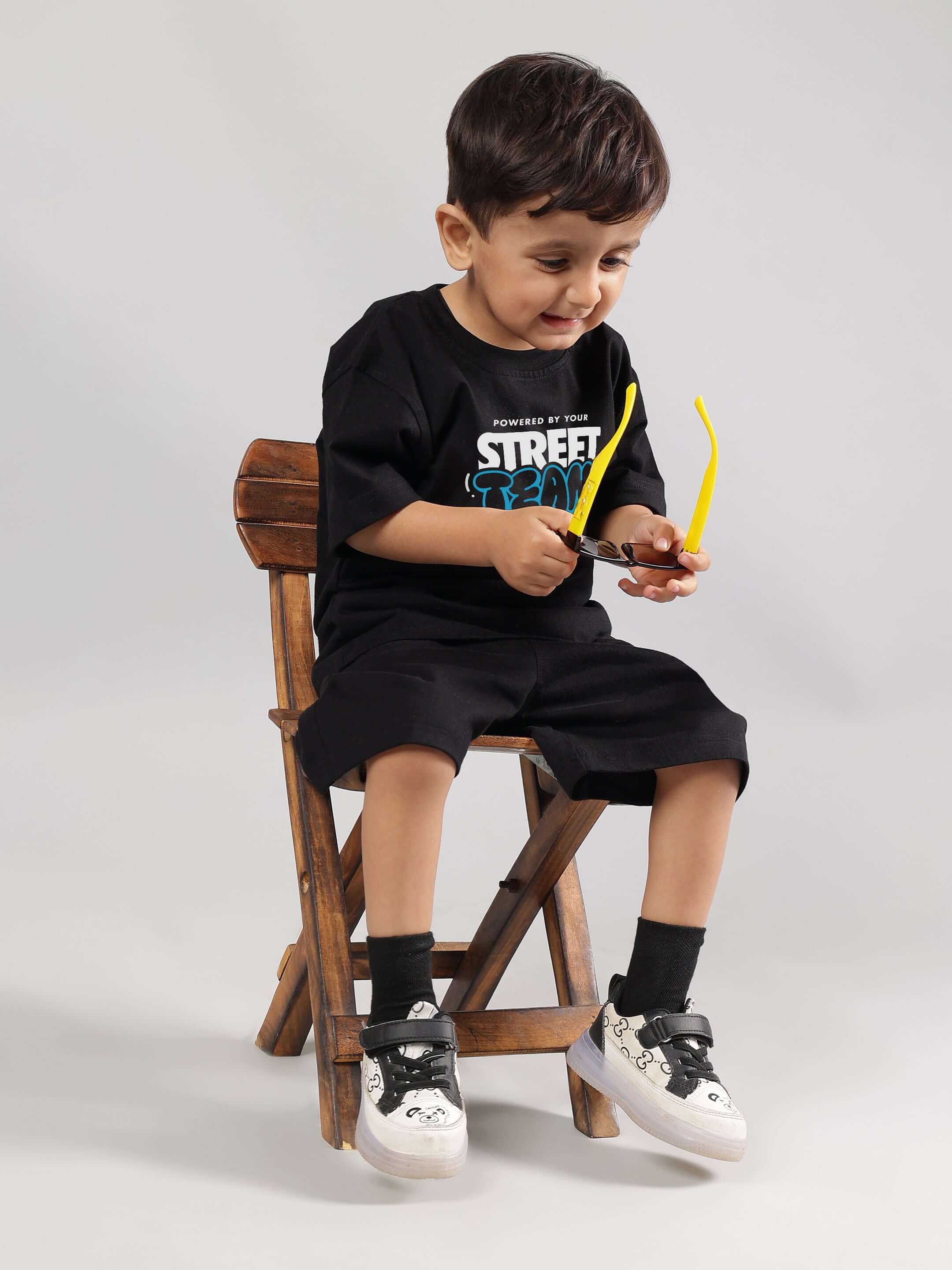 Young boy wearing a black Street Team t-shirt with matching shorts, sitting on a wooden chair and smiling with sunglasses in hand.