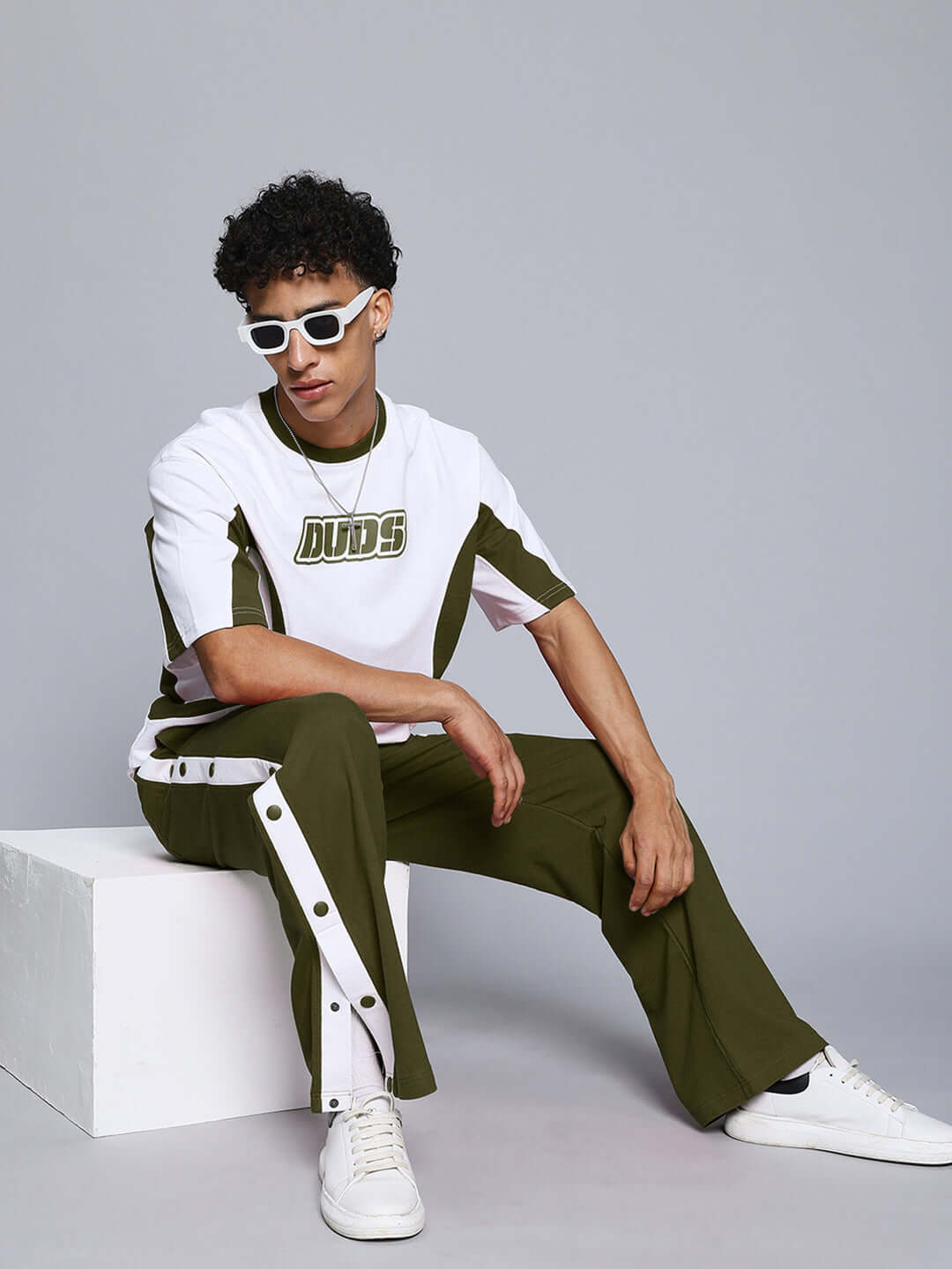Model wearing olive green tracksuit with white accents, sitting on a white block, featuring sporty style and casual design.