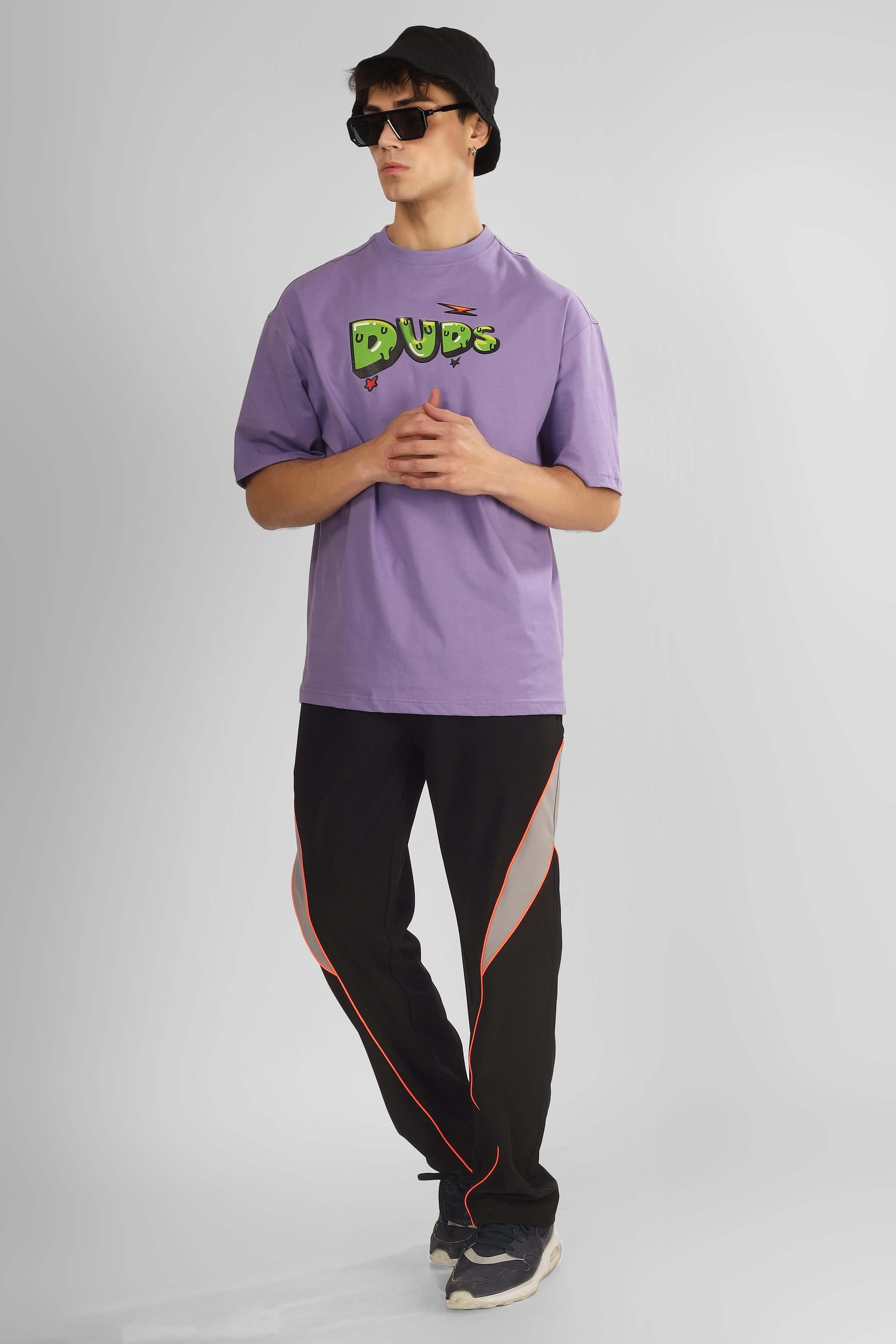 Model wearing a purple Duds t-shirt and black pants with red accents, accessorized with a black hat and sunglasses, in a relaxed pose.