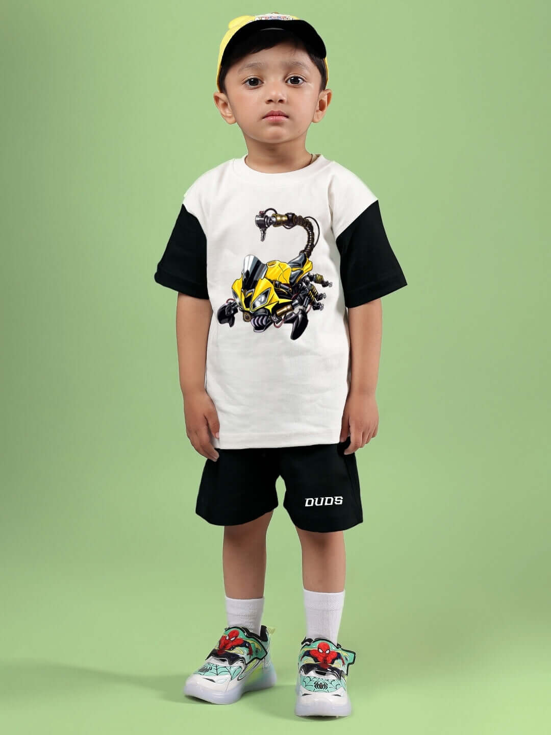 Child wearing STYLISH ROBOT CO-ORD FOR BOYS & GIRLS OFF WHITE BLACK, featuring a robot-print t-shirt and black shorts, on a green background.