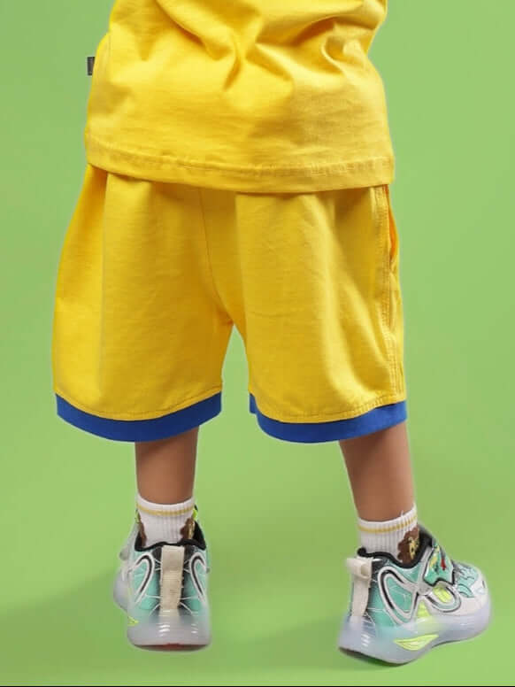 Child wearing SUPER HERO SHORTS FOR BOYS & GIRLS YELLOW on a green background, showcasing vibrant summer fashion.