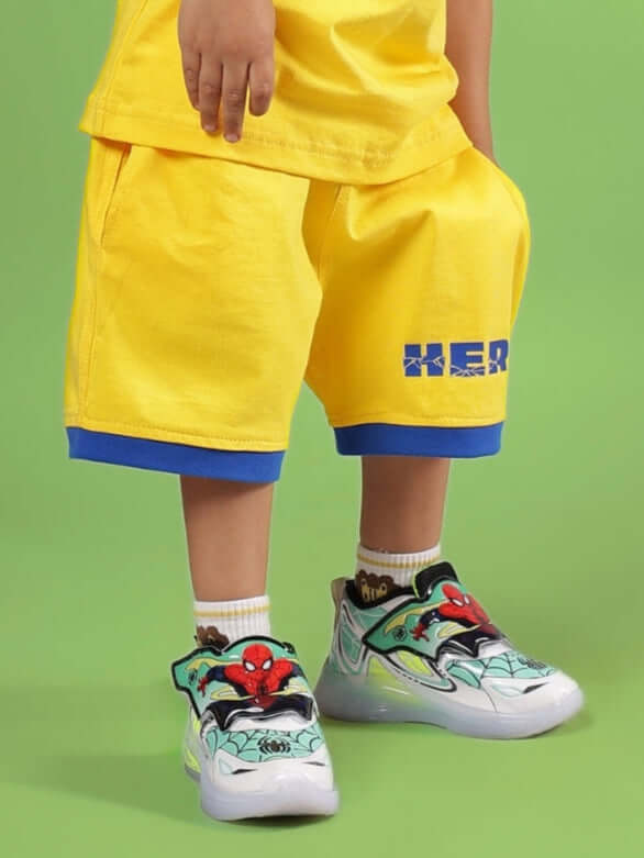 Child wearing SUPER HERO SHORTS FOR BOYS & GIRLS YELLOW with graphic sneakers on green background.